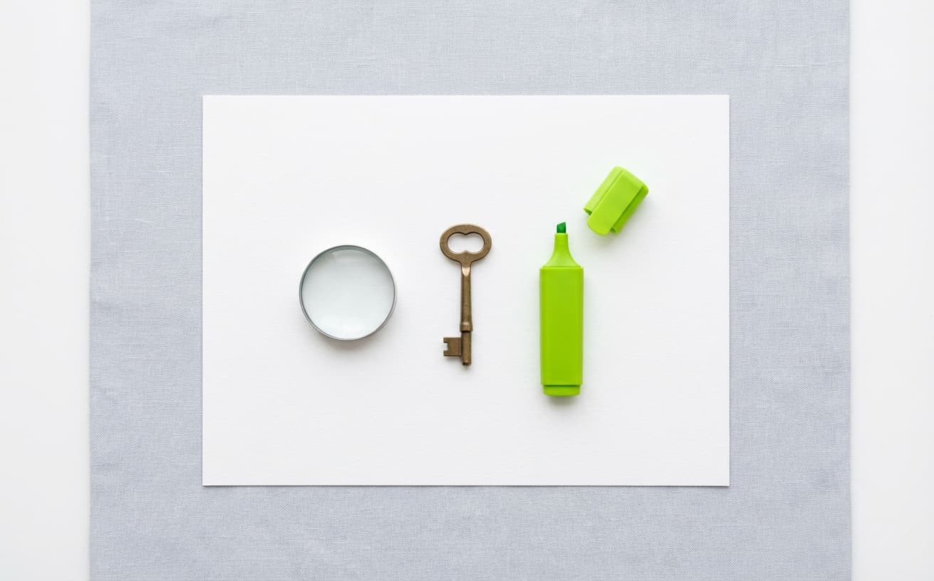 Flat-lay of a white paper with a magnifying glass, brass key, and green highlighter arranged in a row on a light grey linen surface.