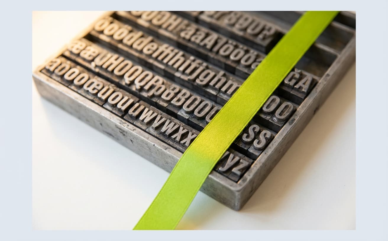Close-up of a metal letterpress typesetting tray with lead characters in neat rows and a green ribbon bookmark across the corner.