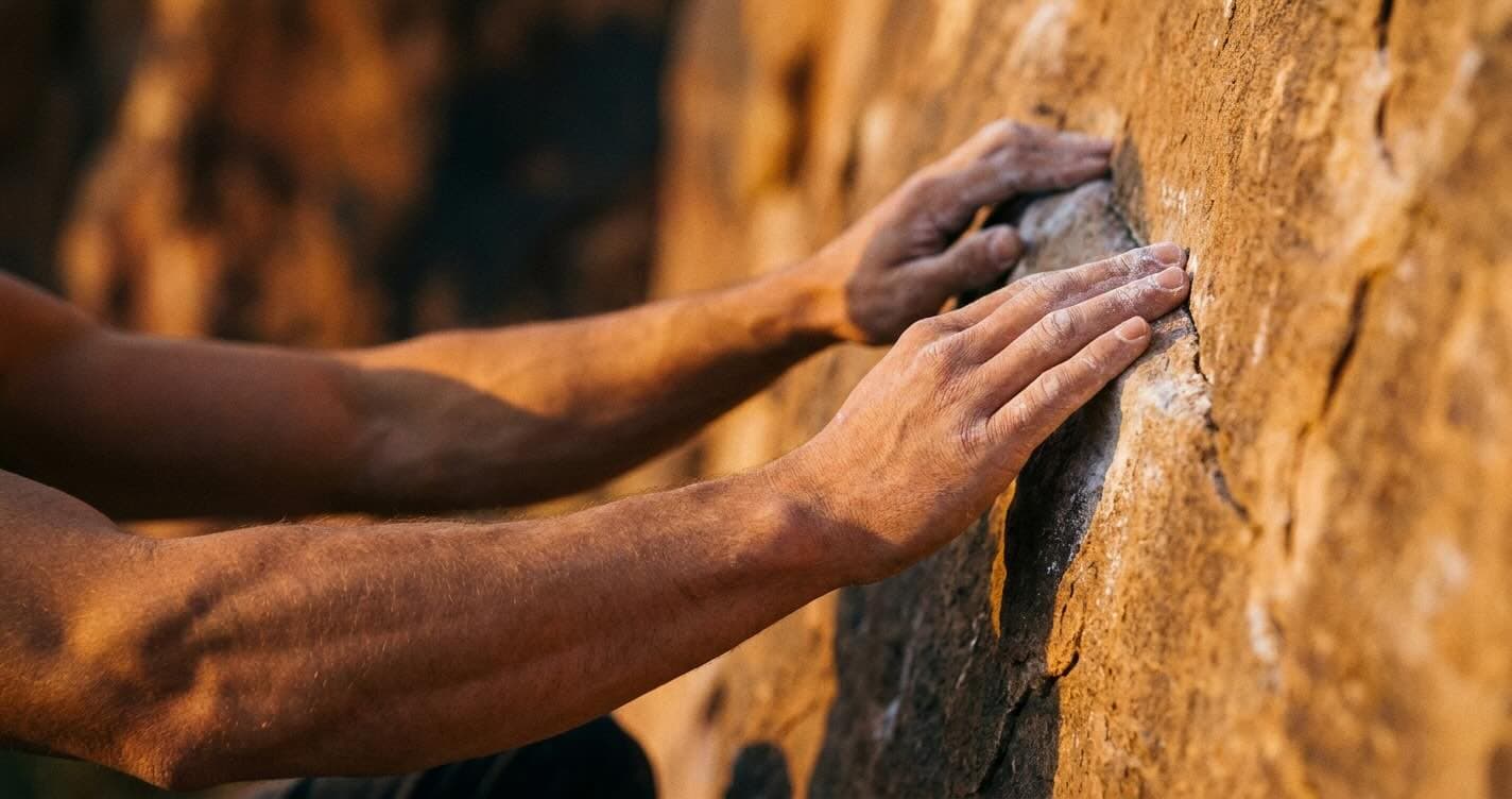 Close-up of chalk-dusted hands gripping a sandstone rock face at golden hour, muscles in the forearm flexed with controlled effort.